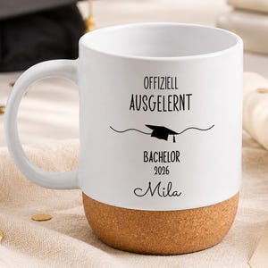 Könnte beinhalten: Ein weißer Keramikbecher mit Korkboden, bedruckt mit dem schwarzen Text Offiziell Ausgelernt, Bachelor 2026 und dem Namen Mila, ergänzt durch eine kleine Grafik einer Doktorhut-Kopfbedeckung mittig zwischen den Textzeilen.