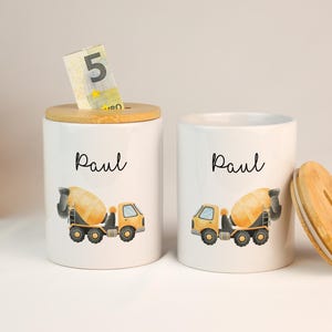 Könnte beinhalten: Zwei weiße Keramik-Sparschweine mit Holzdeckeln. Jede Bank zeigt eine gelbe und graue Betonmischer-Illustration und den Namen "Paul" in schwarzer Schrift. Eine Bank hat einen 5-Euro-Schein eingelegt.
