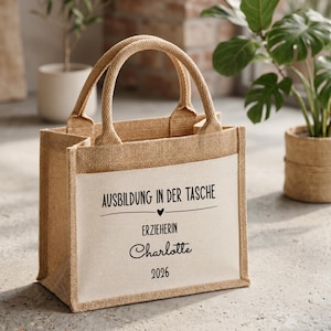 Könnte beinhalten: Eine beigefarbene Jute-Tragetasche mit zwei stabilen Henkeln und einer weißen, rechteckigen Frontpartie. Auf der Vorderseite ist in schwarzer Schrift AUSBILDUNG IN DER TASCHE, ein kleines Herzsymbol, das Wort ERZIEHERIN, der Name Charlotte und 2026 gedruckt.