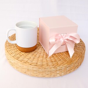 Könnte beinhalten: Ein weißer Keramikbecher mit einem Boden aus Naturkork steht neben einer quadratischen, hellrosa Geschenkbox. Die Box hat eine glatte, matte Oberfläche und ist mit einer breiten, hellrosa Satinschleife an der Vorderseite des Behälters verziert.