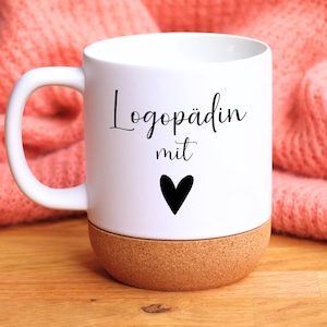 Könnte beinhalten: Eine weiße Keramiktasse mit einem abgerundeten Korkboden. Auf der Vorderseite der Tasse befindet sich der schwarze Schriftzug Logopädin mit, gefolgt von einem schwarzen Herzsymbol. Die Tasse hat einen glatten Henkel an der linken Seite.