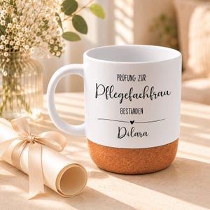 Könnte beinhalten: Ein weißer Keramikbecher mit Korkboden, bedruckt mit dem schwarzen Text PRÜFUNG ZUR Pflegefachfrau BESTANDEN, einem kleinen Herz und dem Namen Dilara, begleitet von einer mit beigem Band gebundenen, gerollten Papierrolle.