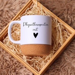Könnte beinhalten: Weißer Keramikbecher mit Korkboden, mit dem Text "Physiotherapeutin mit" und einem schwarzen Herz. Der Becher wird in einer Holzkiste mit Stroh präsentiert.