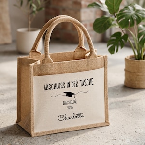 Könnte beinhalten: Eine beigefarbene Jute-Tragetasche mit einer weißen Frontpartie, die den schwarzen Schriftzug ABSCHLUSS IN DER TASCHE, ein Doktorhut-Symbol, das Wort BACHELOR, das Jahr 2026 und den Namen Charlotte in Schreibschrift zeigt.