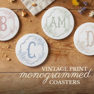 May include: Four white ceramic coasters with floral designs and monogrammed letters B, AM, C, and D. The coasters are arranged on a wooden surface with the text "VINTAGE PRINT monogrammed COASTERS".