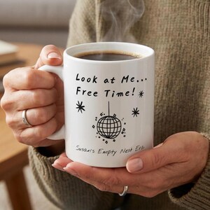 May include: White ceramic mug filled with dark liquid, possibly coffee, held by a person. The mug features the text "Look at Me... Free Time!" with a disco ball graphic and the words "Susan's Empty Nest Era."