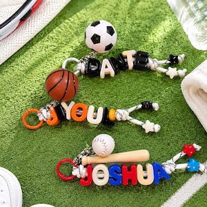May include: Three sports-themed keychains featuring colourful letter beads spelling DANTE, YOU, and JOSHUA. The keychains include miniature football, basketball, and baseball equipment charms, attached to white cord with star-shaped beads at the ends.