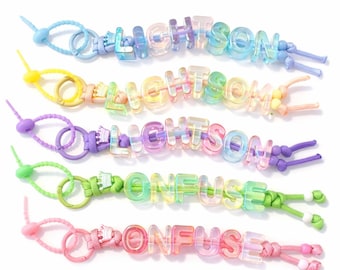 Puede incluir: Cinco pulseras coloridas que deletrean "LIGHTSON" y "CONFUSE" en letras transparentes e iridiscentes. Cada pulsera presenta una combinación de colores diferente, incluyendo azul, amarillo, morado, verde y rosa. Las pulseras están atadas con cordones de colores a juego.
