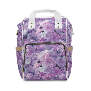 Peut inclure: Un sac à dos avec un imprimé intégral de roses rose pâle et violettes. Le sac comprend une poignée supérieure blanche, des bordures blanches sur les côtés, une poche frontale zippée et des poches latérales. Motif floral sur l'extérieur.