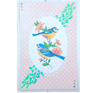 May include: A pink card with white polka dots and a white oval frame with two blue birds and pink flowers. The frame is decorated with a white lace design. There are green leaves in the corners of the card.