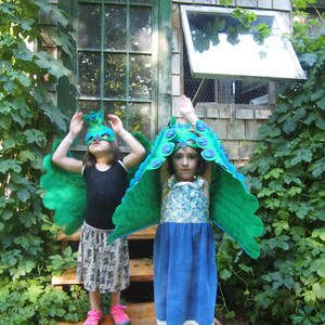 May include: Two children wearing peacock costumes with green and blue wings and masks. The children are standing in front of a wooden building with a window.