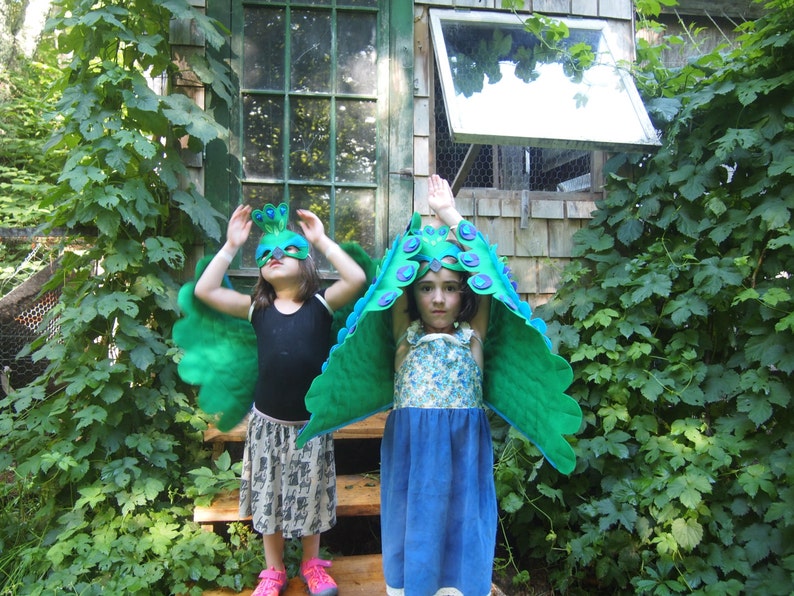 May include: Two children wearing peacock costumes with green and blue wings and masks. The children are standing in front of a wooden building with a window.