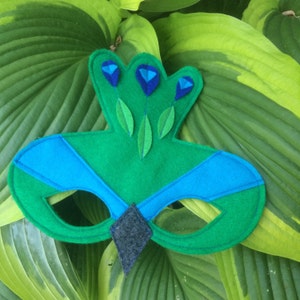May include: A green felt peacock mask with blue accents and a gray felt beak. The mask has eye holes and is designed for children's play.