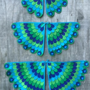 May include: Three pairs of peacock wings made of felt. The wings are in different sizes and are layered on top of each other. The wings are blue, green, and teal.