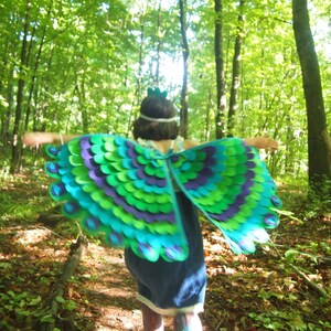 May include: A child wearing a peacock costume with green, blue, and purple feathers runs through a forest. The costume has a crown on top of the head.