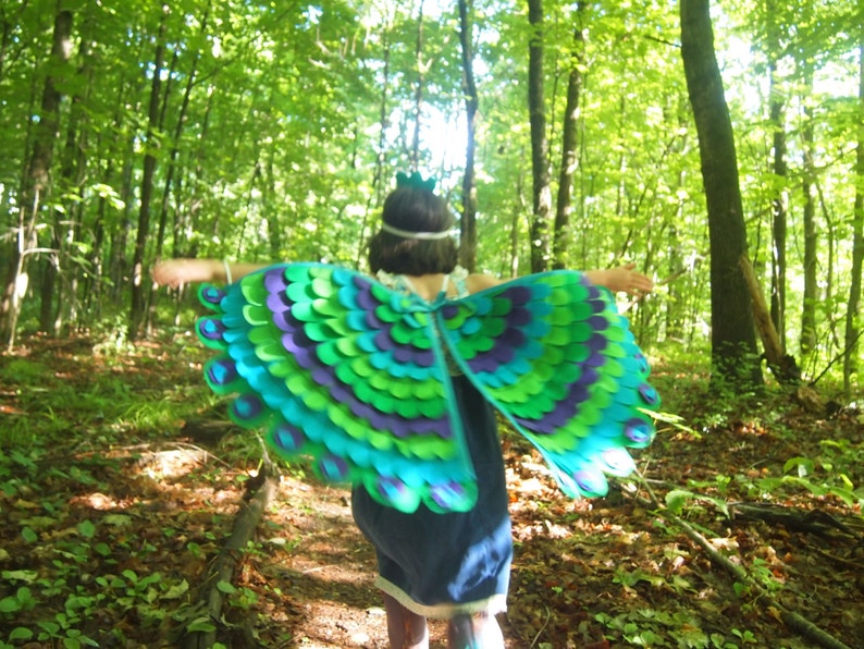 May include: A child wearing a peacock costume with green, blue, and purple feathers runs through a forest. The costume has a crown on top of the head.