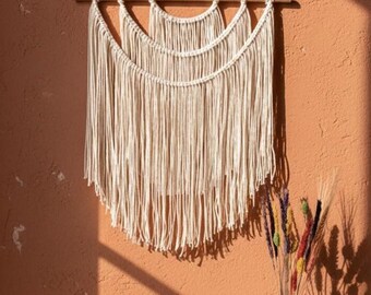 May include: Cream-colored macrame wall hanging with a layered design, suspended from a wooden dowel. The textured piece is displayed against a textured orange wall, with a vase of colorful dried flowers in the foreground.