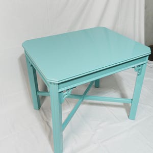 May include: A square, turquoise table with a glossy finish. The table has a decorative apron and legs with cross-bracing. The table's dimensions are approximately 76 cm long, 76 cm wide, and 46 cm high.