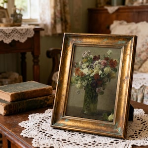 May include: A framed floral print with a gold-colored, distressed frame. The artwork depicts a bouquet of flowers in a clear vase. The frame is propped up on a wooden table with a lace doily, with antique books in the background.