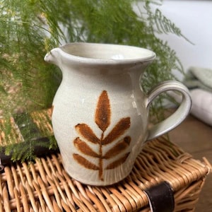 May include: A cream-colored ceramic pitcher featuring a brown leaf pattern on the front. The pitcher has a rounded body, a wide spout, and a curved handle on the side. The surface has a smooth, glazed finish with visible earthy tones.