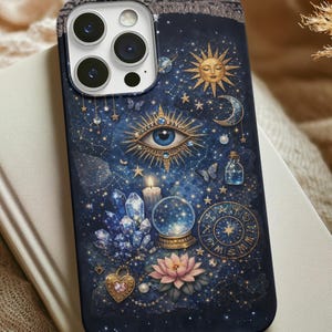 May include: A dark blue phone case featuring a celestial design with an eye, sun, moon, stars, crystals, and a snow globe. The case is resting on a book and a beige fabric.