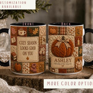 May include: Two ceramic mugs with black handles and rims featuring a patchwork quilt design in autumn colors. The front mug displays the text 'COZY SEASON LOOKS GOOD ON YOU' with icons of books, leaves, and a mug. The back mug features a pumpkin and the text 'ASHLEY Officially in her cozy era'.