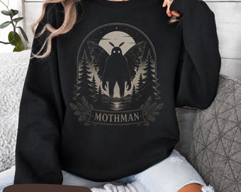 Puede incluir: Una sudadera negra de manga larga con un estampado gráfico de una criatura alada con ojos brillantes de pie en un bosque bajo una luna llena. El diseño está encerrado en un marco arqueado con el texto MOTHMAN impreso en una cinta inferior.