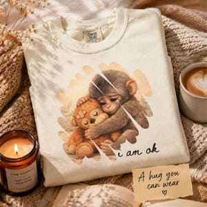 May include: Cream-colored sweatshirt with a graphic of two hugging monkeys and the text "i am ok". The sweatshirt also has the text "A hug you can wear" on a small card. A lit candle and a cup of coffee are also in the image.