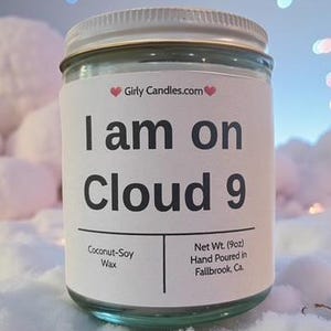 May include: A clear glass jar candle with a silver lid, labeled "I am on Cloud 9." The label also includes "GirlyCandles.com" and "Coconut-Soy Wax." The candle is set on a fluffy, white surface with a blurred background of lights.