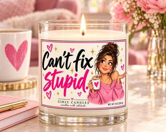 May include: A clear glass candle with a white wax fill and a lit wick. The label features a cartoon of a person in a pink sweater holding a mug, with the text "Can't fix Stupid." and "GIRLY CANDLES candles with attitude" and "NET WT. 9 OZ (255 g)".
