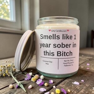 May include: A clear glass jar candle with a white label that reads "Smells like 1 year sober in this Bitch." The candle is on a wooden surface with dried flowers and petals. The candle is 9oz.