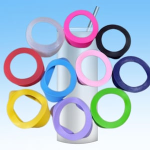 Puede incluir: Una colección de anillos de silicona coloridos en varios tonos, incluyendo rosa, azul, verde y amarillo, dispuestos alrededor de un vaso blanco con una pajita plateada. Los anillos parecen estar diseñados para encajar en la base del vaso.