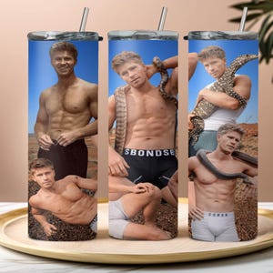 May include: A stainless steel tumbler featuring a collage of images of a man with snakes and lizards. The images are set against a blue sky and desert landscape. The tumbler has a silver lid and a straw.