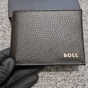 May include: A black leather wallet with a textured finish, displaying the gold-coloured "BOSS" logo. The wallet is presented with its matching black box, also bearing the "BOSS" logo in white.