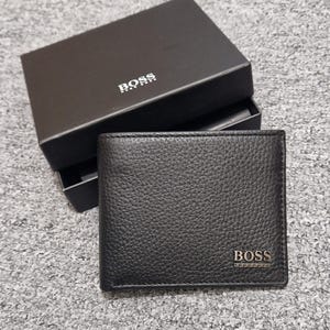 May include: A black leather wallet with the "BOSS" brand name embossed on the front. The wallet is square and has a textured surface. A black box with the brand name is behind the wallet.
