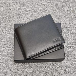 May include: A black leather wallet with the embossed "BOSS" logo, presented on a matching black box. The wallet features a classic bi-fold design with visible stitching. The overall aesthetic is sleek and minimalist.