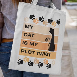 May include: A cream tote bag with a black cat silhouette and the text "MY CAT IS MY PLOT TWIST" in black and orange. The bag features paw prints in black and tan.