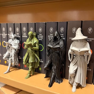 May include: Four figurines of characters from a fantasy novel, each holding a book. The figures are in silver, olive green, black, and gray. The figurines are displayed in front of a row of books with the title "The Lord of the Rings".
