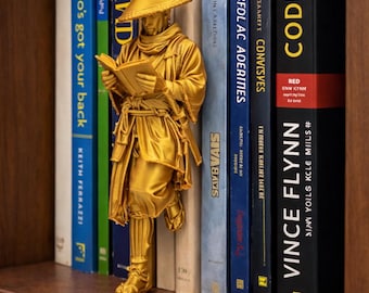 May include: A gold-colored figurine of a monk reading, positioned between books on a shelf. The books have titles like "Who's Got Your Back" and "Code Red". The figurine is a decorative item for book lovers.