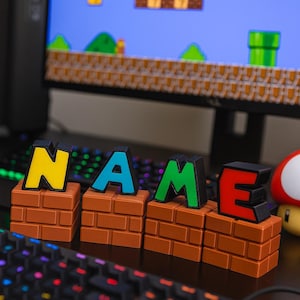 Può includere: Una colorata esposizione con la parola "NAME" in lettere vivaci sopra strutture a forma di mattoni. Le lettere sono gialle, blu, verdi e rosse. Un monitor di computer mostra una scena di videogioco classico sullo sfondo.