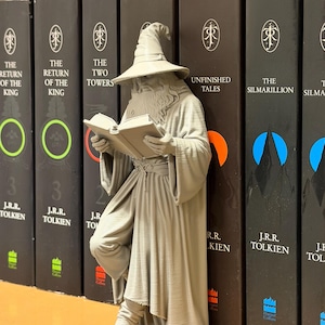 May include: A gray wizard figurine reading a book, leaning against a row of black books. The books have titles like "The Return of the King" and "The Two Towers" by J.R.R. Tolkien. The figurine is detailed, with a hat and robe.