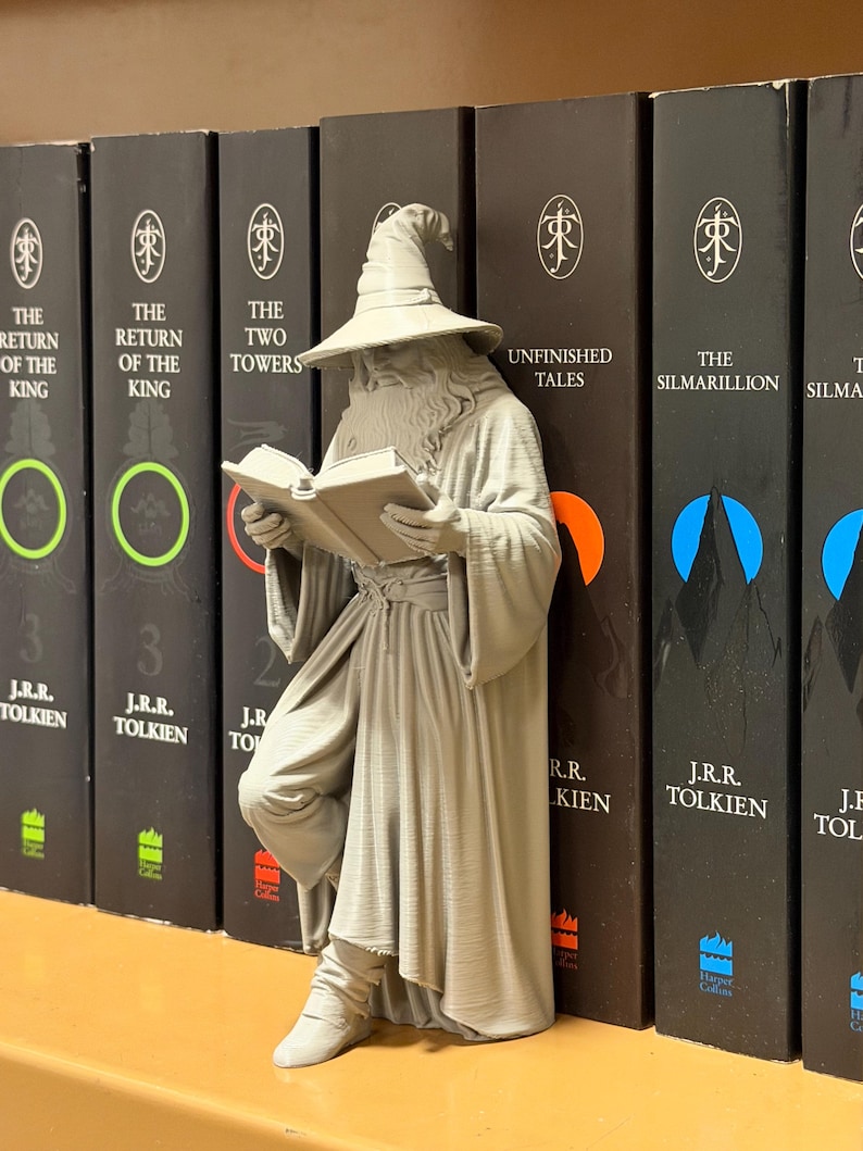May include: A gray wizard figurine reading a book, leaning against a row of black books. The books have titles like "The Return of the King" and "The Two Towers" by J.R.R. Tolkien. The figurine is detailed, with a hat and robe.