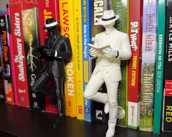 May include: Two figurines of a person in a suit and fedora hat, one black and one white, are leaning against a shelf of colorful books. The figurines are reading books. The books have titles like "Jenny Lawson" and "John Scalzi".