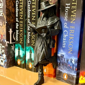 May include: A black plague doctor figurine reading a book, positioned against a row of books. The books are titled "Gardens of the Moon" and "House of Chains" by Steven Erikson. The figurine features a hat, beak mask, and long coat, with intricate detailing.
