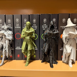 May include: Four figurines of characters from The Lord of the Rings, each holding a book. The figures are silver, green, black, and gray, and stand in front of a row of books with titles from the series.