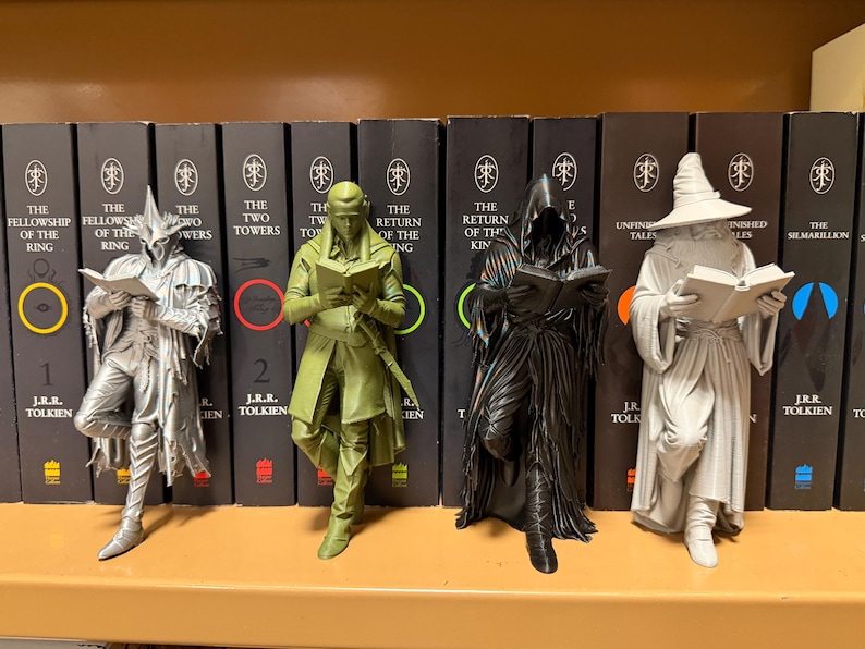 May include: Four figurines of characters from The Lord of the Rings, each holding a book. The figures are silver, green, black, and gray, and stand in front of a row of books with titles from the series.