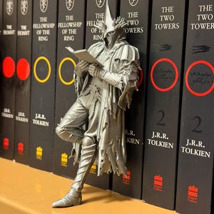 May include: A silver-colored figurine of a character from The Lord of the Rings, leaning against a row of books. The figurine is reading a book. The books are black with white text and feature the titles of the series. The figurine is approximately 6 inches tall.