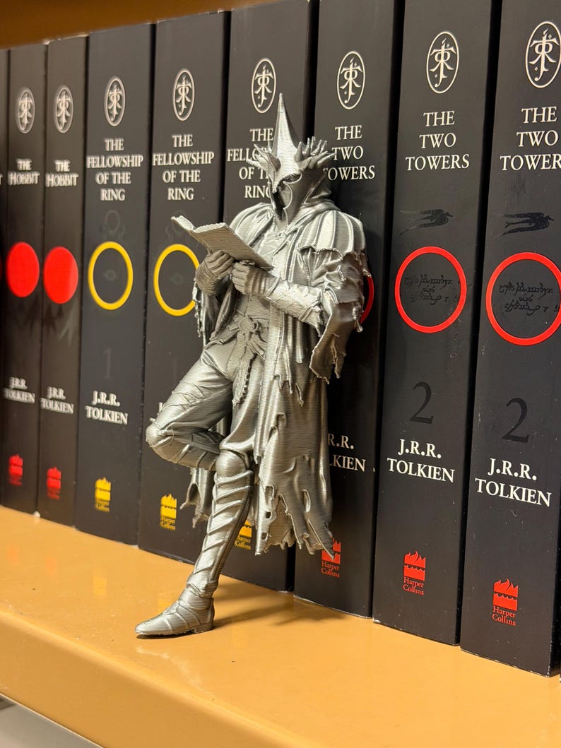 May include: A silver-colored figurine of a character from The Lord of the Rings, leaning against a row of books. The figurine is reading a book. The books are black with white text and feature the titles of the series. The figurine is approximately 6 inches tall.