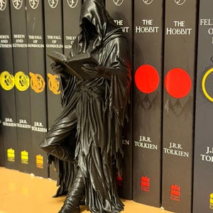 May include: A black figurine of a cloaked figure reading a book, leaning against a row of books. The books have black covers with white text and a circular logo. The figurine has a detailed, textured appearance.