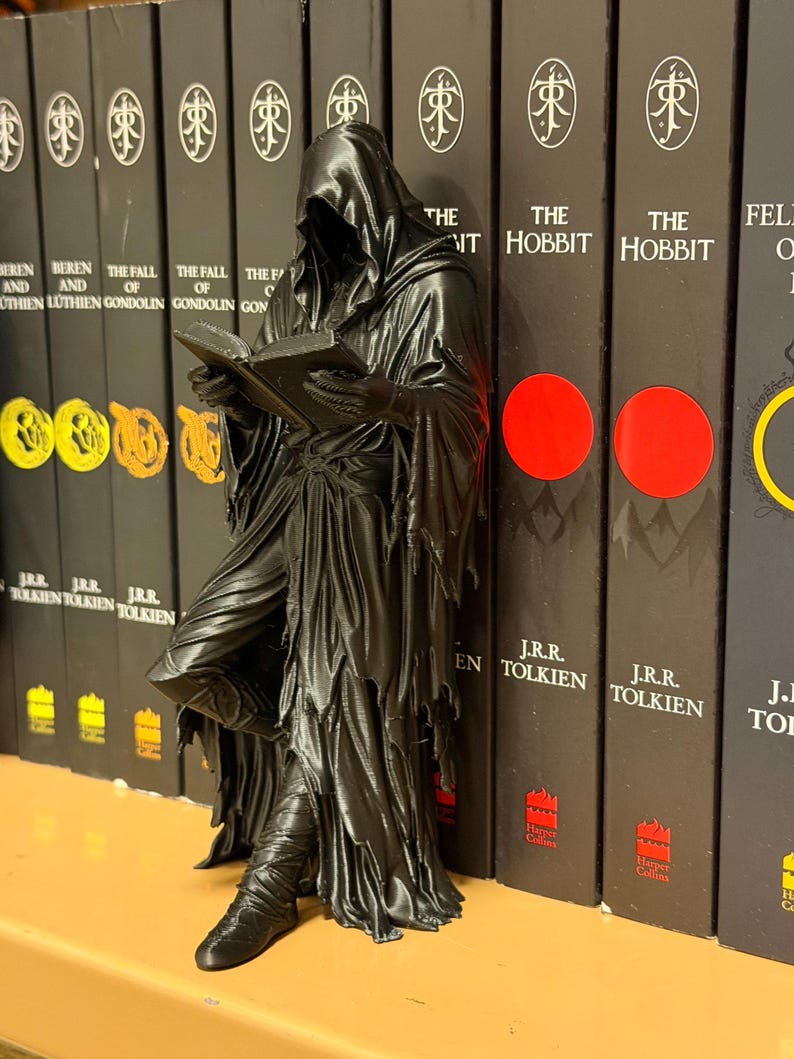 May include: A black figurine of a cloaked figure reading a book, leaning against a row of books. The books have black covers with white text and a circular logo. The figurine has a detailed, textured appearance.